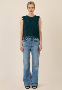 Mujer de pie vistiendo una blusa sin mangas de encaje verde oscuro, jeans de tiro alto y pierna ancha en azul claro, y sandalias negras abiertas en los dedos.