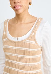 Stripete strikket tanktop i beige over en hvit teksturert skjorte med lange ermer, med rund hals og dekorativ knapp.