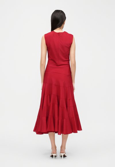 Γυναίκα με μακριά σκούρα μαλλιά που φοράει κόκκινο midi φόρεμα χωρίς μανίκια με τουρλουμπούκι φούστες και λευκές ψηλές γόβες, στέκεται γυρισμένη πλάτη σε λευκό φόντο.