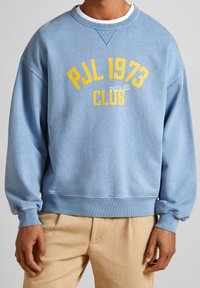 Světle modrý sweatshirt s kulatým výstřihem vyrobený z bavlny, zdobený žlutým potiskem s nápisem "PJL 1973 CLUB PHYS. ED." a elastickými manžetami a lemem.