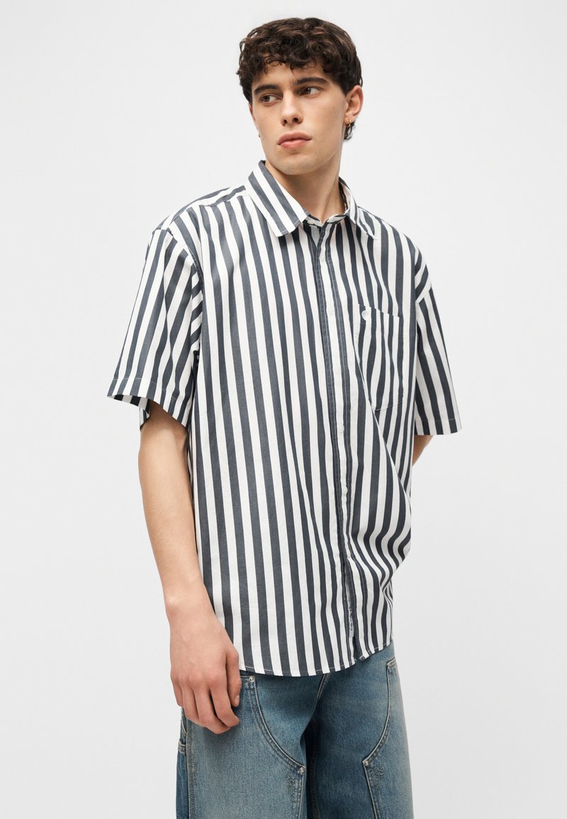 Giovane uomo che indossa una camicia a maniche corte con bottoni e righe verticali bianche e nere e jeans di denim blu, che guarda di lato.