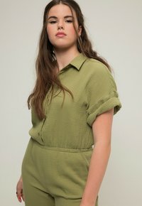Olivgrüner, strukturierten Stoff-Jumpsuit mit Kragen, kurzen umgeschlagenen Ärmeln und elastischem Taillenbund. Verfügt über Knöpfe vorne.