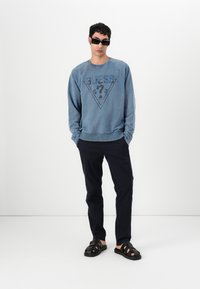Lys blå hettegenser med en mørk grafisk logo, kombinert med mørke bukser og svarte sandaler. Designet har rund hals og ribbede mansjetter.