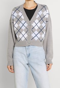 Harmaa lyhythihainen neuletakki, jossa on napit, valko-mustan vinoneliökuvio ja ribbireunat. Täytetään mustan paidan ja vaaleiden farkkujen yli.