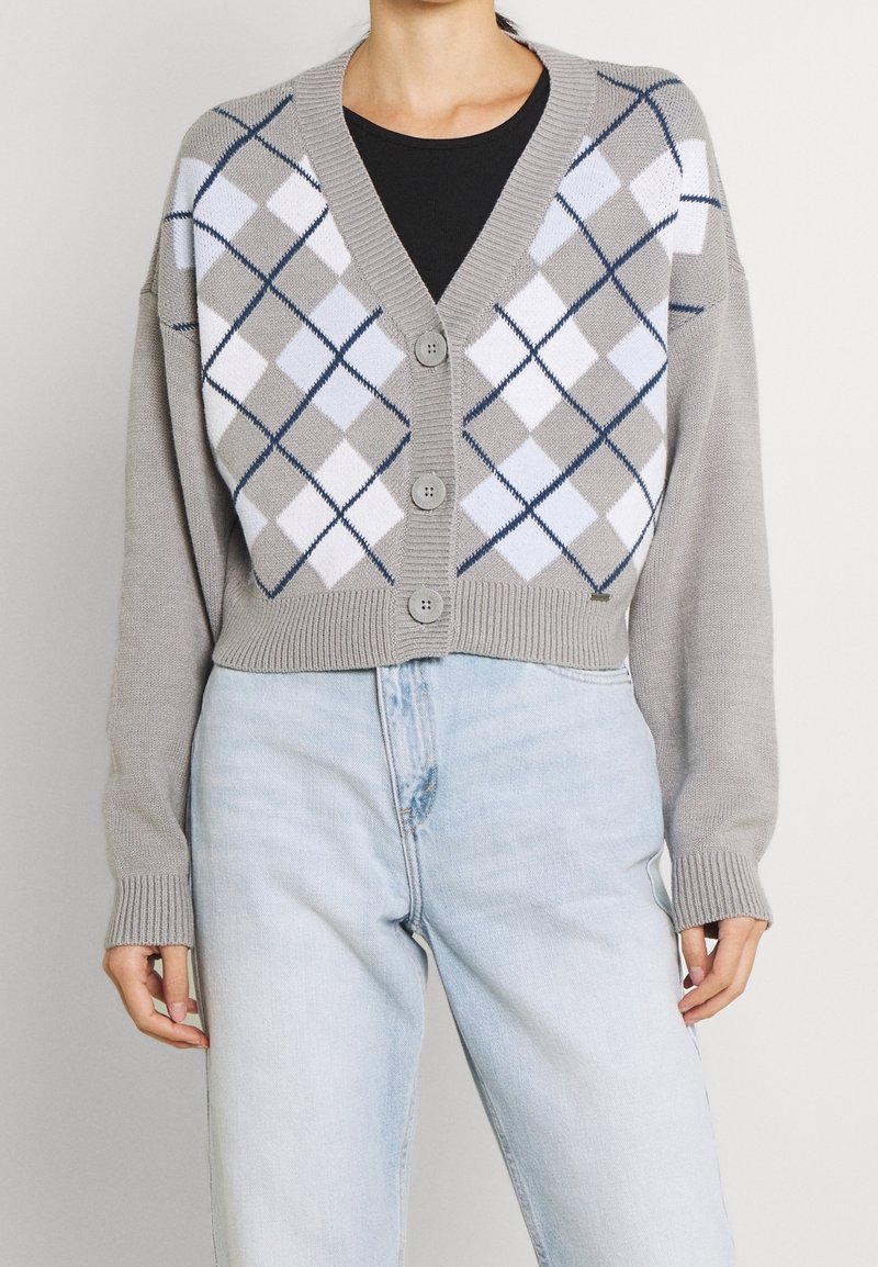 Harmaa lyhythihainen neuletakki, jossa on napit, valko-mustan vinoneliökuvio ja ribbireunat. Täytetään mustan paidan ja vaaleiden farkkujen yli.