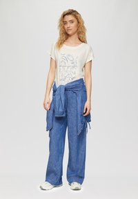 Hellbeige Kurzarm-T-Shirt mit einem strukturierten blauen Grafikdesign und Text, kombiniert mit lockeren blauen Jogginghosen mit Kordelzug und einem gebundenen blauen Pullover.