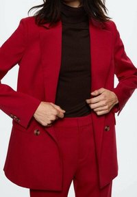 Červený upravený blazer vyrobený z hladké látky, s dvouřadým designem, hnědými knoflíky a klopou s zářezy, kombinovaný s hnědým rolákem.