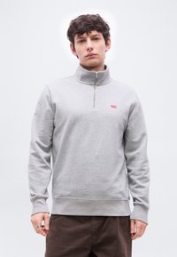 Νέος άνδρας που φοράει ανοιχτό γκρι sweatshirt με μισό φερμουάρ και κόκκινο λογότυπο Levi's, συνδυασμένο με σκούρο καφέ παντελόνι, στέκεται μπροστά σε ένα απλό φόντο.