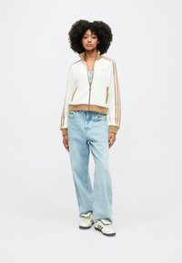 Chaqueta blanca de cremallera con detalles acanalados marrones y tres rayas marrones en las mangas, combinada con jeans de pierna ancha azul claro y zapatillas blancas.