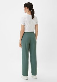 Green wide-leg trousers with a smooth texture and elastic waistband, paired with a white short-sleeve shirt, viewed from the back.