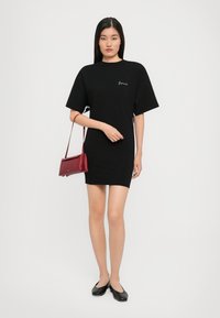 Γυναίκα στέκεται φορώντας ένα μαύρο μίνι φόρεμα με κοντό μανίκι, μαύρες φλατ παπούτσι και κρατώντας μια μικρή κόκκινη τσάντα στον ώμο της, μπροστά από ουδέτερο φόντο.