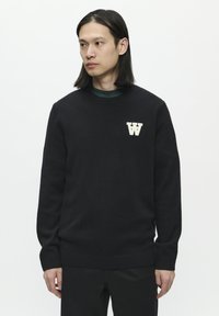 Schwarzer Strickpullover mit gerippten Bündchen und Saum, mit einem cremefarbenen "W"-Logo auf der Brust. Glatte Textur mit Rundhalsausschnitt.