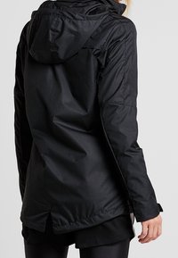 Zwarte waterdichte jas met een capuchon, met een taille die aangesnoerd kan worden, lange mouwen en een iets langere achterkant voor extra bedekking. Textuurstofafwerking.