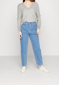 Pull gris à col en V avec manches longues, associé à un jean en denim bleu clair taille haute. Le mannequin porte des baskets blanches. Tenue simple et décontractée.
