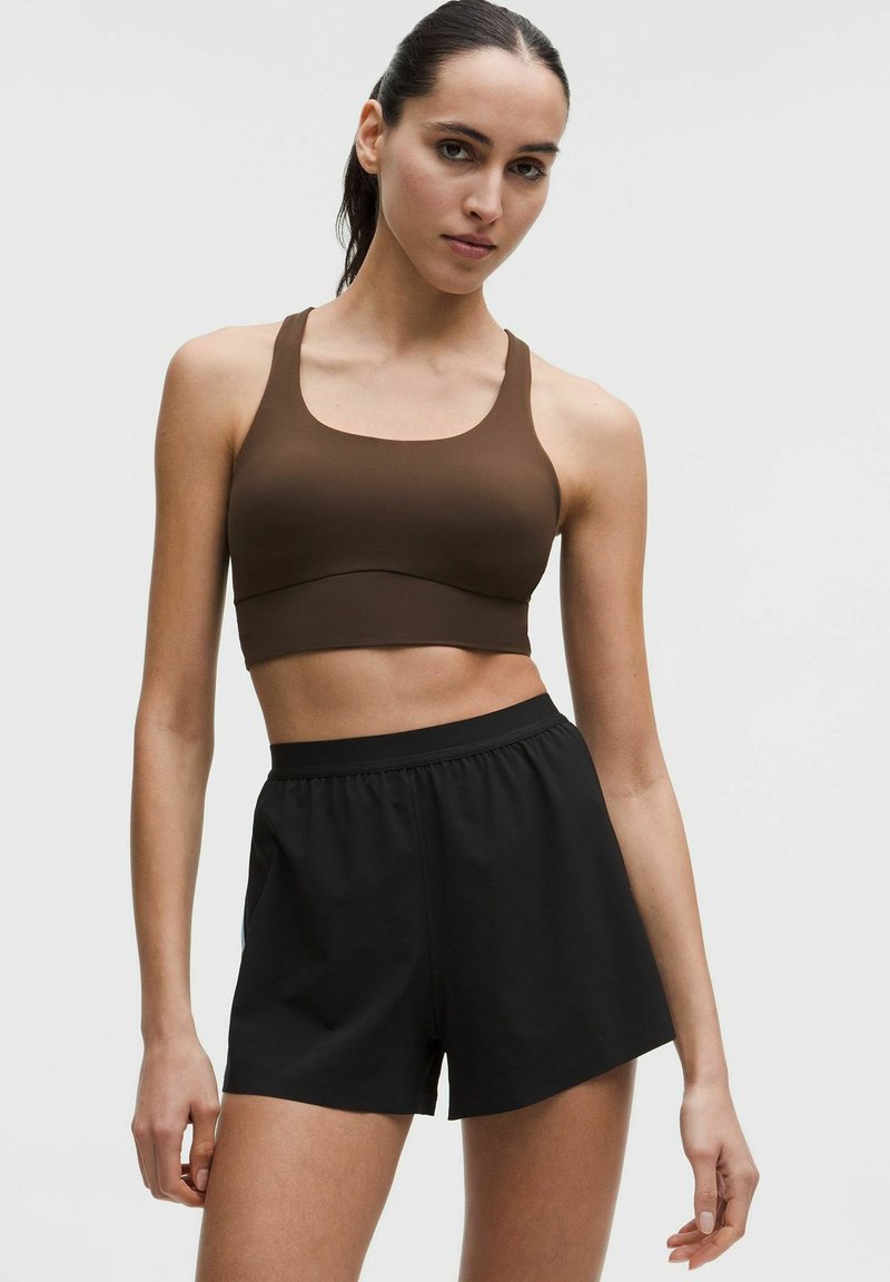 Žena s tamnom kosom vezanom u rep, odjevena u smeđi sportski grudnjak i široke crne sportske hlače, stoji na jednobojnoj pozadini.