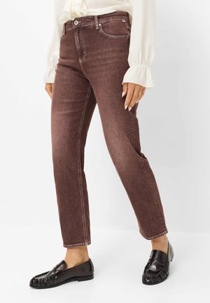 Femme portant un jean droit marron foncé, une blouse blanche à manches longues avec des poignets à volants, et des mocassins en cuir noir, debout sur fond blanc.