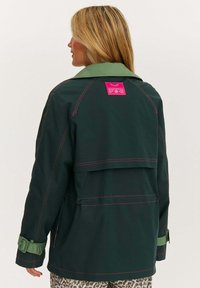 Tmavě zelená bunda se světle zeleným límcem, růžovým švem a jasně růžovou etiketou na zadní straně. Má přezkové rukávy a volný střih.