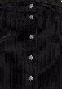 Falda de pana negra con estrías verticales y cinco botones a presión metálicos en la parte delantera, combinada con una blusa de canalé verde oscuro.