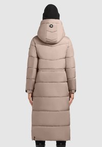 Hosszú, bélelt kabát világos bézs színben, kapucnival, steppelt mintázattal és fekete fém kiegészítőkkel. A szövet sima és texturált hatású.
