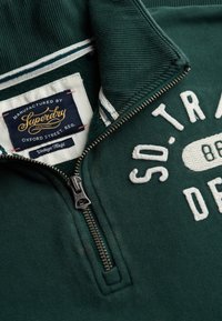 Felpa verde con collo con zip, caratterizzata da un testo ricamato bianco. Il tessuto ha una trama morbida e all'interno è visibile un'etichetta con il marchio.