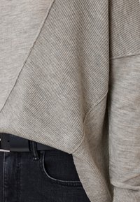 Maglione di colore beige a maglia con una texture a coste, caratterizzato da un design drappeggiato. Indossato sopra jeans di denim scuro, con una cintura nera in evidenza.