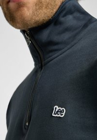 Felpa navy con colletto alto, presenta una cerniera in argento e il logo "Lee" ricamato. Il tessuto è liscio con una finitura leggermente strutturata.