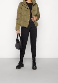 Olivgrön vadderad jacka med dragkedja och tryckknappsförslutning, kombinerad med svarta åtsittande byxor och svarta snörkängor. Svart handväska ingår.