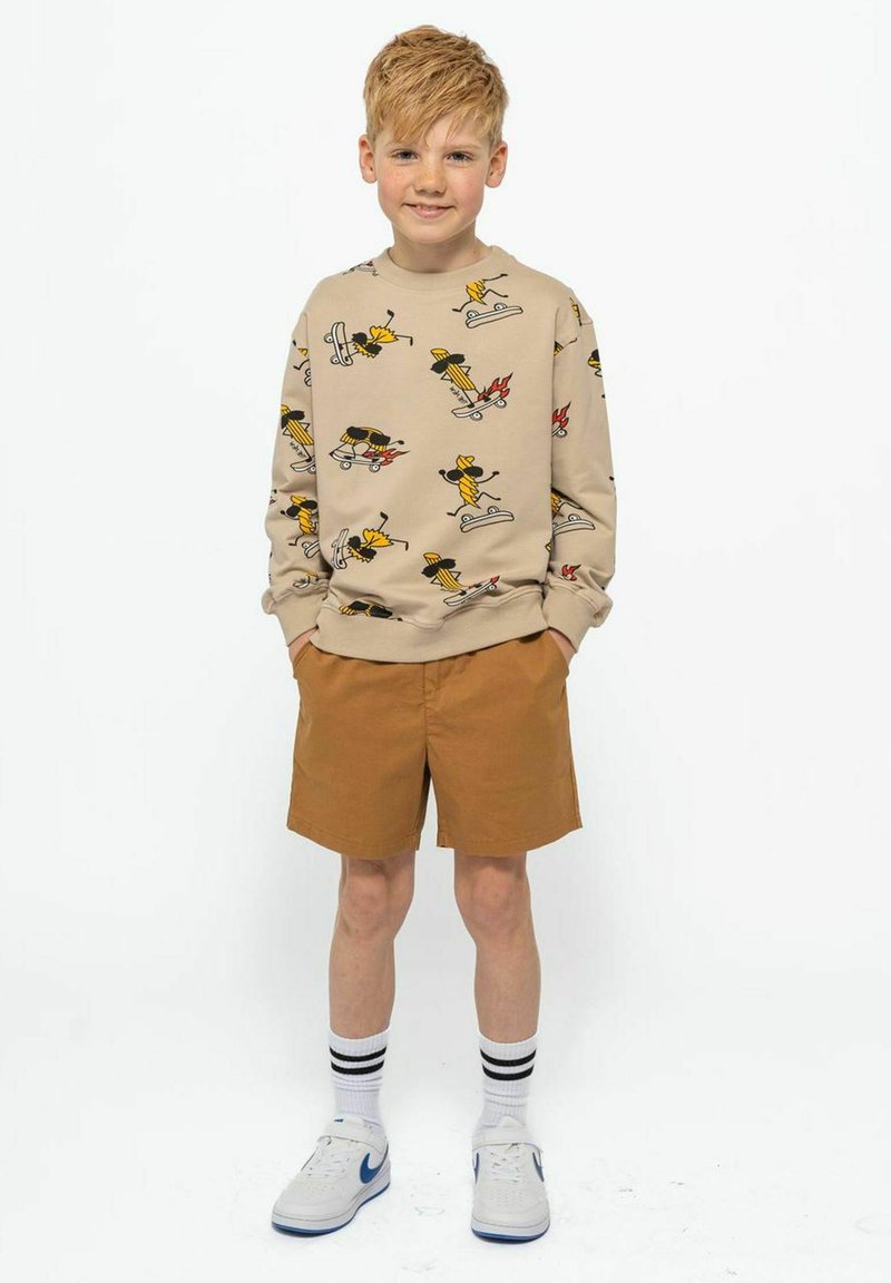 Felpa beige con motivi a cartone animato, pantaloni corti marroni, sneaker bianche con dettagli blu e calzini a righe bianchi e neri.