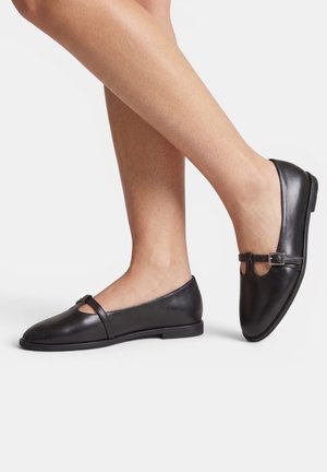Gambe che indossano scarpe basse in pelle nera stile Mary Jane con una piccola fibbia sul collo del piede, mostrate su uno sfondo semplice.