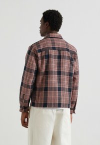 Koszula w kratę w kolorze pomarańczowym i czarnym o luźnym kroju, z długimi rękawami i rękawami zapinanymi na guziki, wykonana z teksturowanej tkaniny. Wyświetlana z widoku z tyłu.