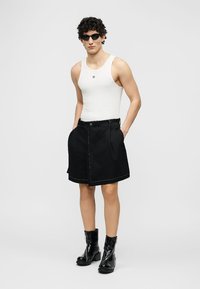 Man wearing black sunglasses, white sleeveless top, black skirt with belt, and shiny black ankle boots, standing with hands in pockets.