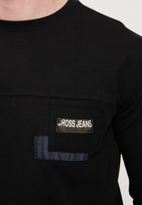 Czarna bawełniana koszulka z krótkim rękawem, z przednią kieszenią naszywaną z denimu. Metka z napisem "CROSS JEANS" na zielonym tle.