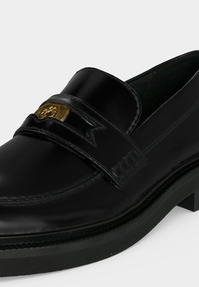 Mocassin en cuir noir avec une semelle épaisse et un détail d'emblème doré sur la bride située sur le dessus de la chaussure.