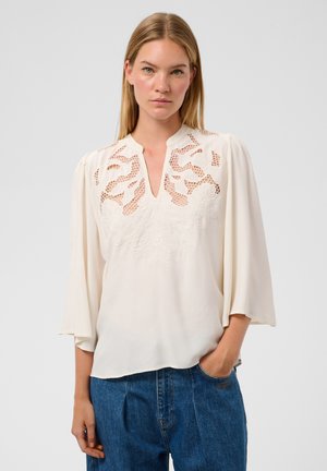 Frau mit langen blonden Haaren, die eine weiße Bluse mit Spitzendetails und blaue Jeans trägt, steht mit einer Hand in der Tasche vor einem schlichten Hintergrund.
