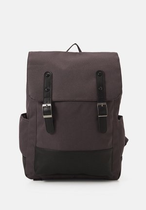 Sac à dos en tissu gris foncé avec rabat, deux sangles en cuir noir avec boucles argentées, poches latérales et une poignée de transport sur le dessus.