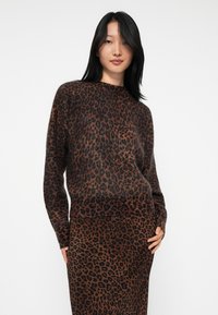 Mulher com cabelo longo e preto, vestindo um conjunto de suéter e saia com padrão de leopardo em marrom e preto, de pé contra um fundo branco liso.