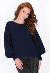 Granatowy sweter z dzianiny o szerokich rękawach i ściągaczu u dołu. Miękka tekstura, casualowy styl. Noszony z ciemnymi dżinsami dla swobodnego wyglądu.