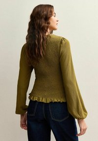 Top smock verde oliva con maniche a sbuffo lunghe, tessuto testurizzato e orlo arricciato; abbinato a jeans blu scuro.