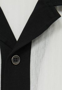 Nava scurtă alb-negru cu guler, cu mânecă scurtă; are un buton circular cu patru orificii și material texturat.