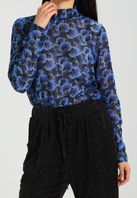 Långärmad blus med ett blått blommönster på ett genomskinligt tyg, med hög halsringning. Matchas med strukturgröna svarta byxor med hög midja.