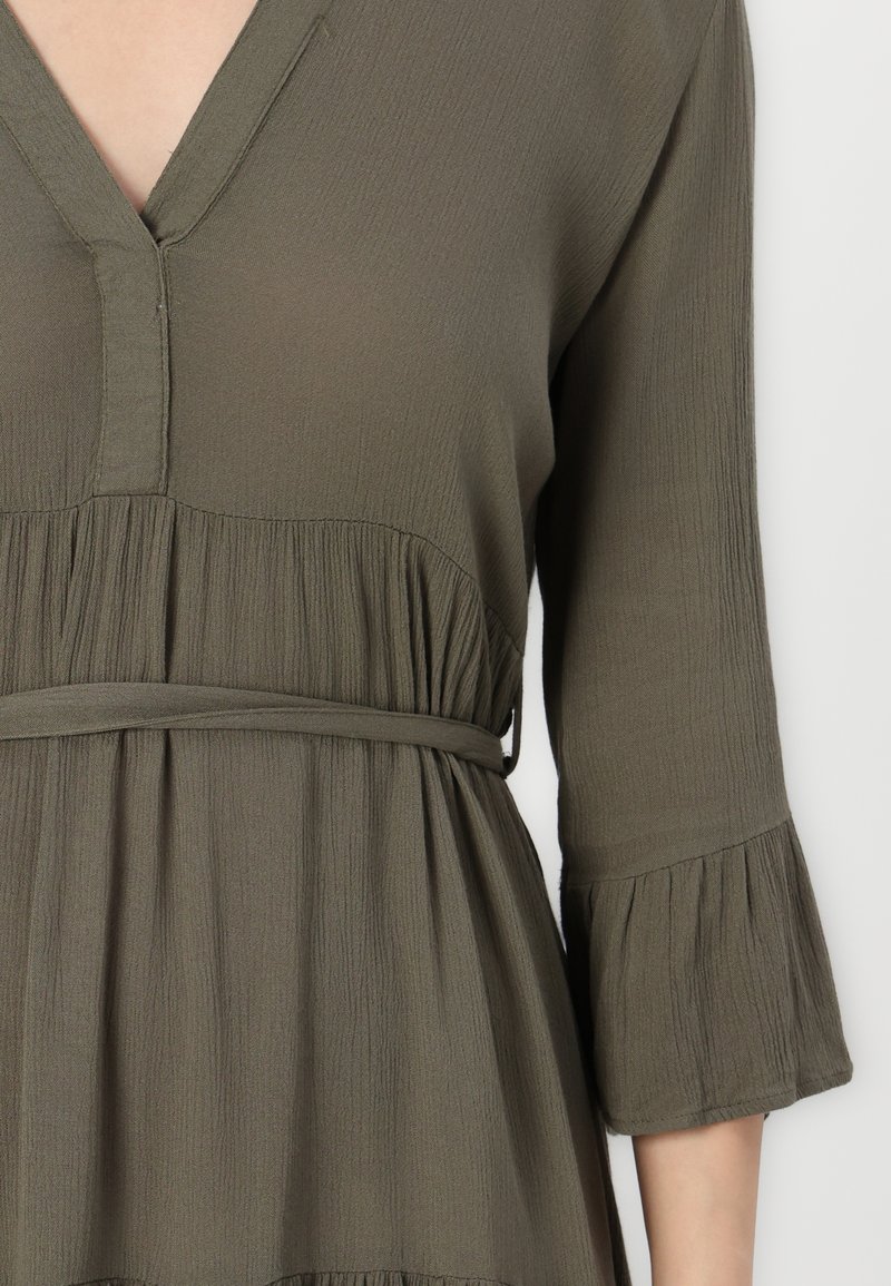 Robe vert olive avec un décolleté en V, manches cloche trois quarts, tissu texturé et taille cintrée. Présente des plis doux au bas de la jupe.