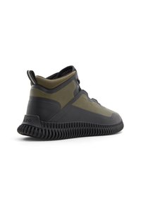 Zapatilla alta en verde oliva y goma negra, con una suela texturizada, forma redondeada y diseño minimalista con cordones prominentes.