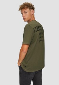 Olivgrünes T-Shirt mit kurzen Ärmeln, das auf der Rückseite mit großen schwarzen Buchstaben bedruckt ist und einen abgerundeten Saum hat. Hergestellt aus weichem Baumwollmaterial.