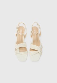 Sandales à talons blanches avec un bout carré, des sangles en tissu texturé nouées à l'avant et une fermeture à boucle sur le dos.