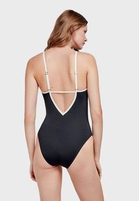 Maillot de bain une pièce noir avec un dos en V profond, des bretelles crème en accent et un tissu lisse. Il présente une coupe ajustée et un matériel minimal.