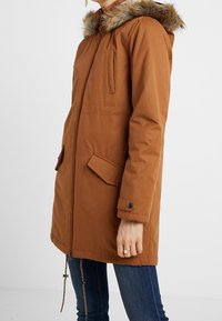 Brown padded coat with faux fur hood, front zip, buttoned pockets, elastic drawstring at the hem, and a smooth, durable texture.