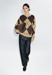 Beżowo-brązowy sweter w wzór argyle z czerwonymi diamentami, zestawiony z ciemnoniebieskimi dżinsami. Modelka trzyma ciemnobrązową trójkątną torbę. Proste tło.