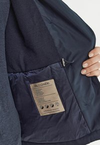 Mörkblå jacka med en slät textil utsida, kashmirinsida och en rektangulär etikett med skötselråd och materialdetaljer.