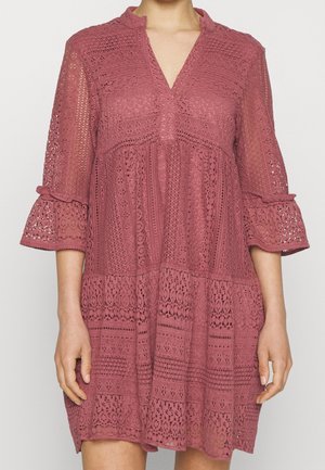 Kobieta nosi różową, koronkową sukienkę do kolan z rękawami 3/4 w kształcie dzwonu i dekoltem w serek, ozdobioną misternymi wzorami kwiatowymi i geometrycznymi.