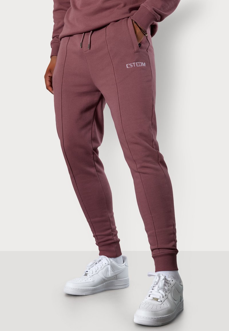 Bordeauxfarbene Jogginghosen mit seitlichen Nähten und elastischen Bündchen, ausgestattet mit Taschen und einem kleinen gestickten Logo. Kombiniert mit weißen Sneakers.